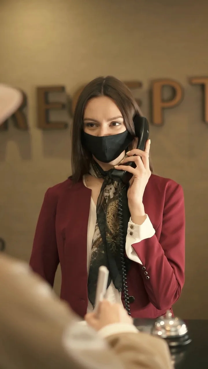A receptionist in a red blazer answers the phone at a hotel reception desk as a guest signs in.
