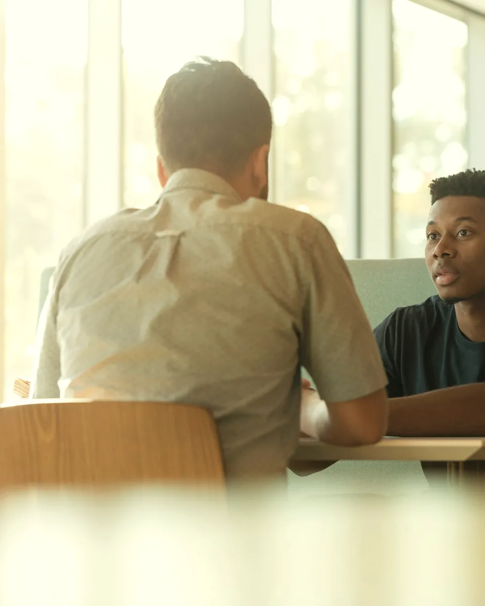 Two people talk across a table in window light, an interview in progress.