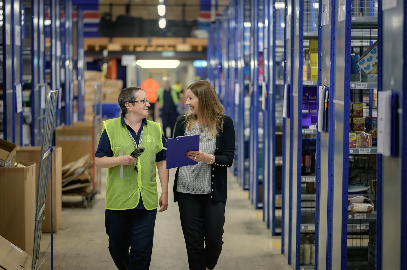 Two colleagues walk a warehouse aisle, reviewing notes.